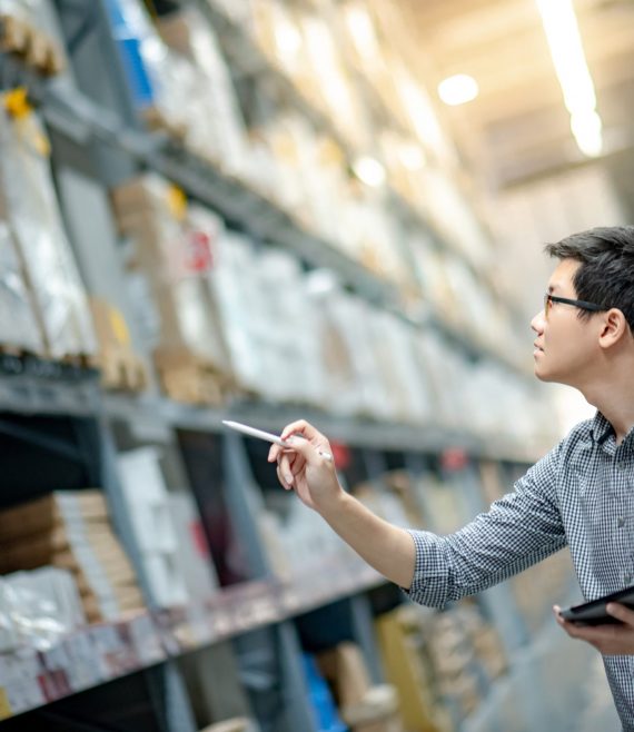 Young Asian man worker doing stocktaking of product in cardboard box on shelves in warehouse by using digital tablet and pen. Physical inventory count concept